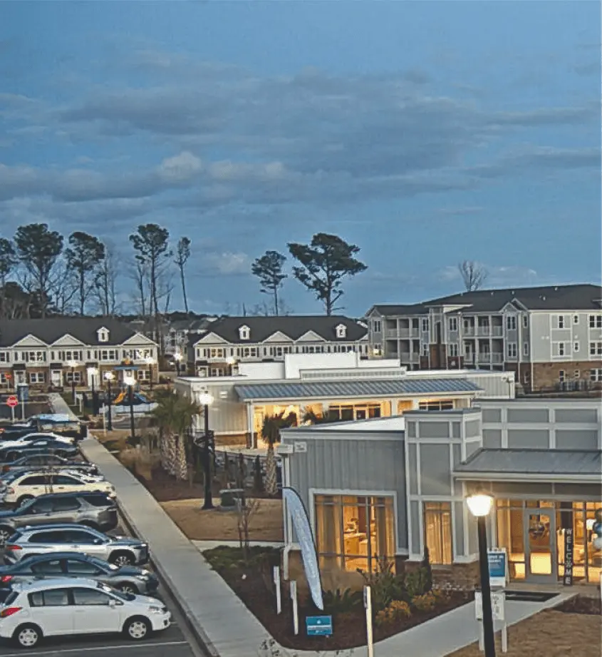  A modern residential complex with multiple buildings, a parking lot filled with cars, and landscaped walkways. The sky is partly cloudy in the background.
        