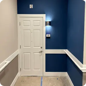  A white door is set into a blue wall, illuminated by a wall sconce above it. The area has white trim and handrails along the walls.